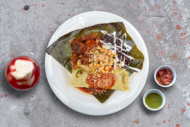 Pork Carnitas Mole Tamal, shown on a table with pork with a organic greens-veggie tamal, ancho chile mole sauce, black beans, chipotle corn salsa, sour cream and cotija cheese.
