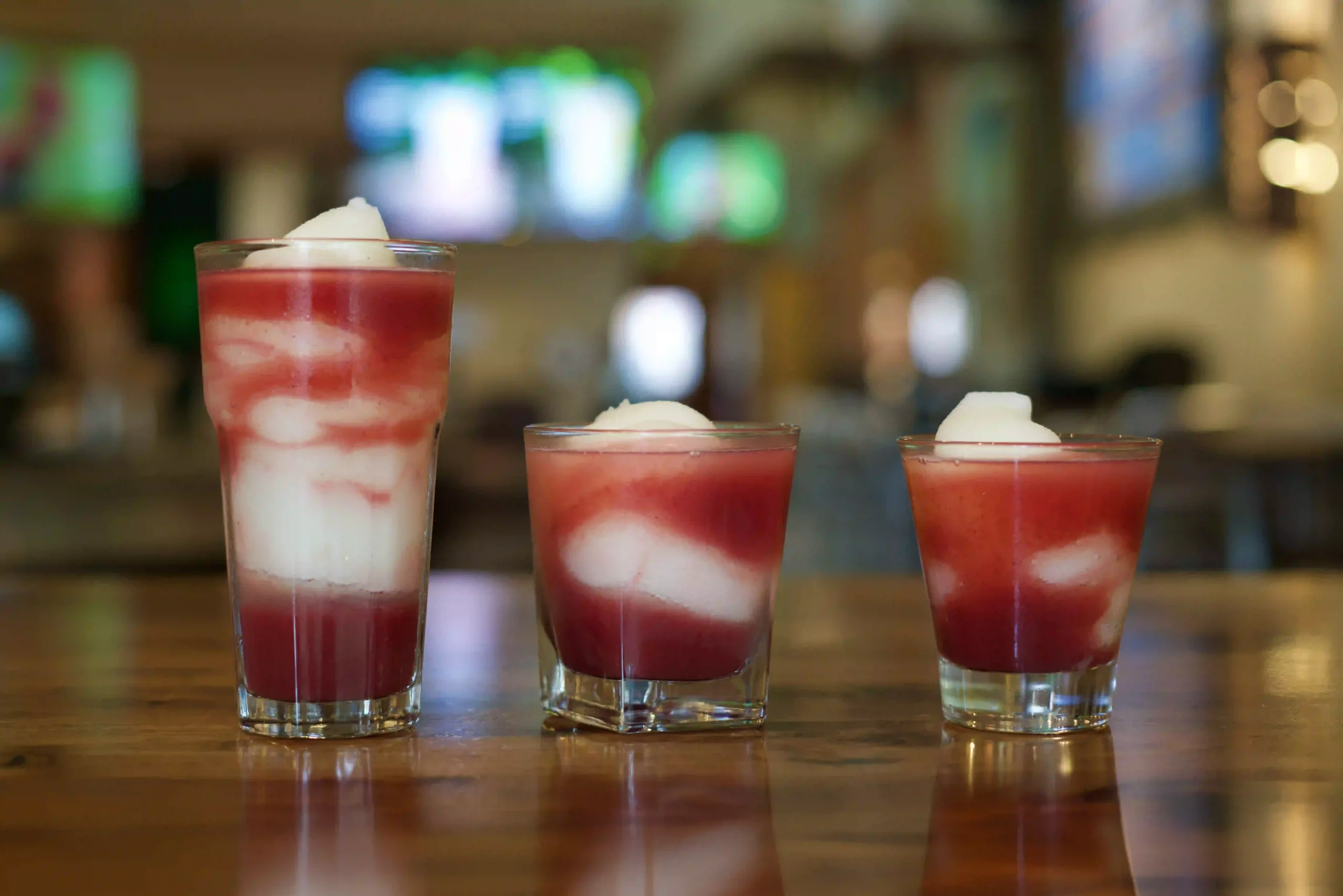 Three glasses with red and white layered drinks topped with frozen margarita on a wooden bar surface.