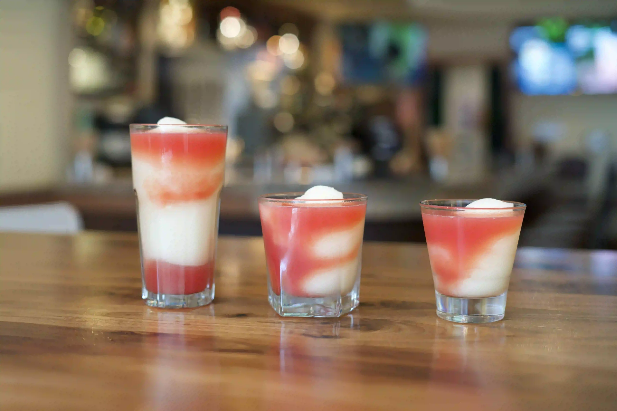 Three layered red-and-white beverages in small clear glasses on a wooden bar surface, with a blurred bar background (foreground focus).