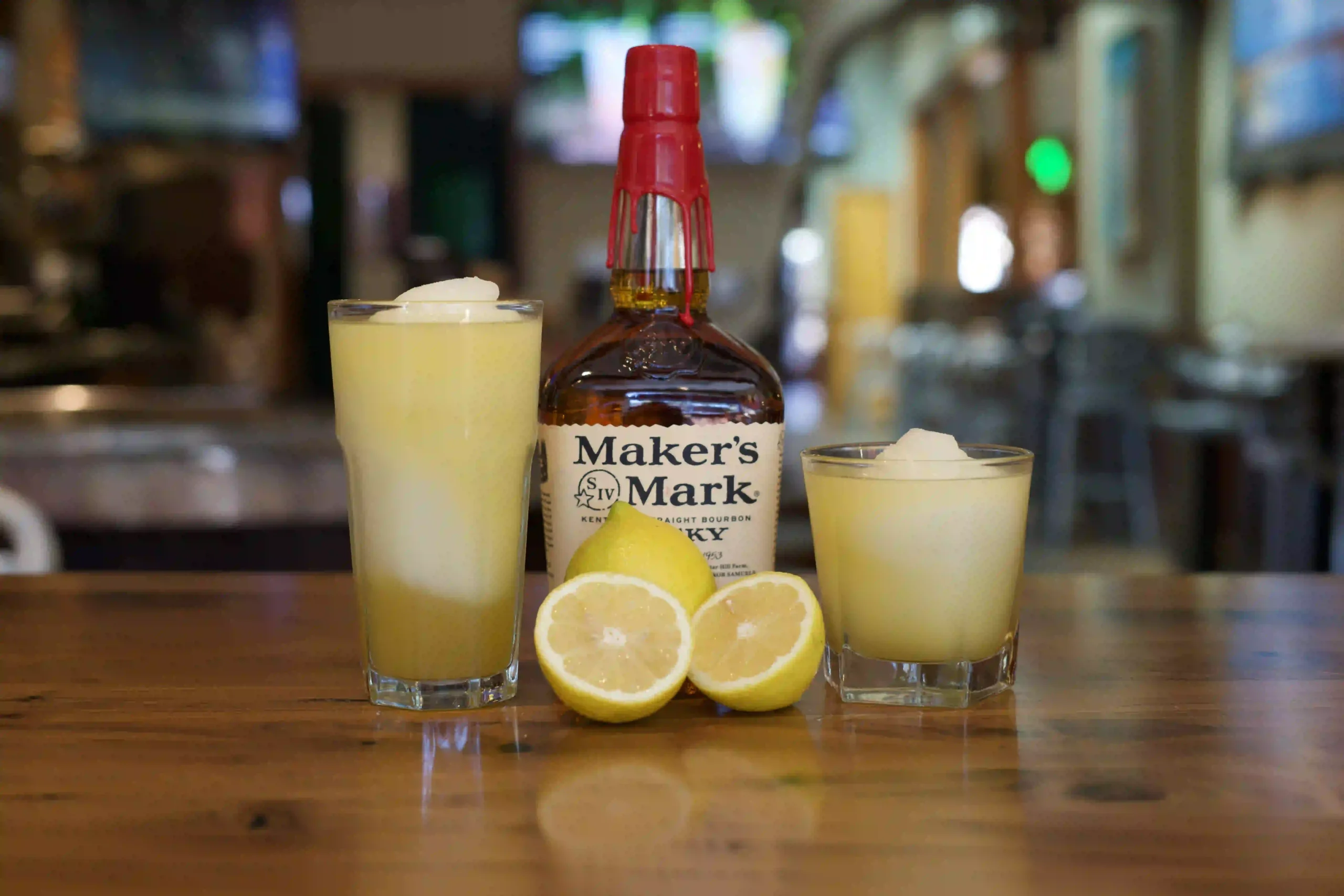 Two yellow cocktails with ice on a wooden bar, centered around a Maker's Mark bourbon bottle and lemon halves in front.